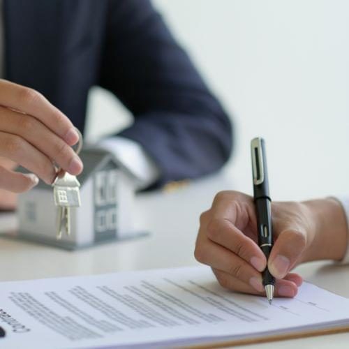 Primer plano de la mano de una persona firmando un contrato junto a otra que tiene en su mano una llave de una propiedad