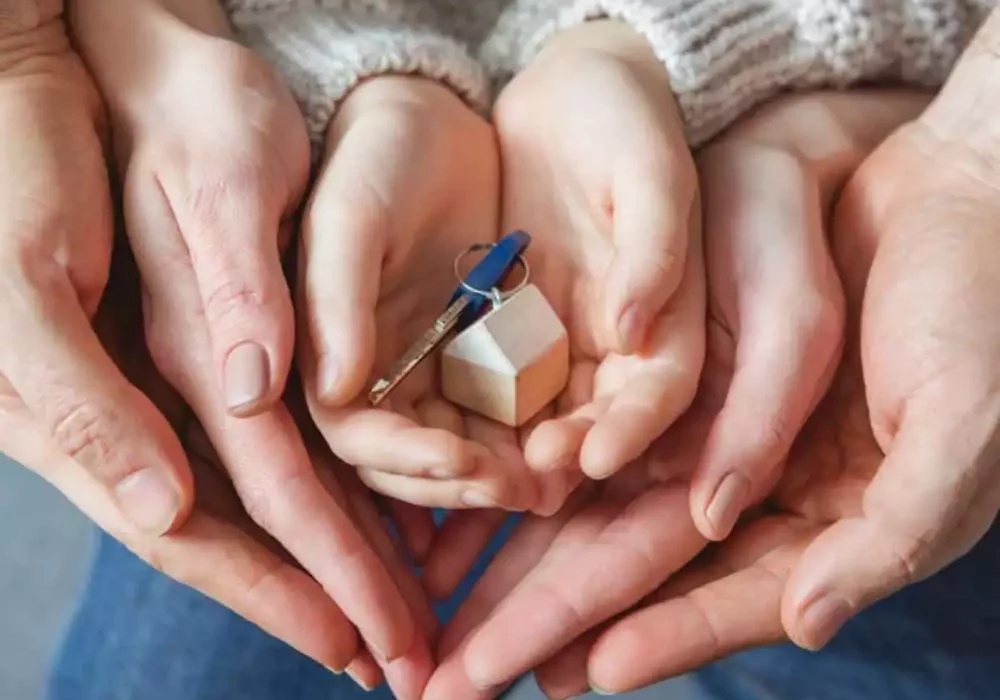 Manos de dos adultos que acogen y envuelven las manos de una niña o niño que entre ellas, sostiene un llavero con forma de casa y una llave